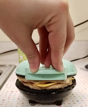 Hand pushing down on lid of waffle iron to grill sandwich